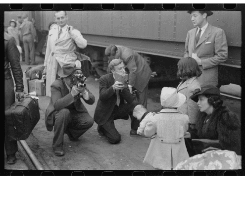 A group of reporters and photographers gather around a woman and two children on a train platform. Two men kneel with cameras, while others stand nearby holding bags and coats. A train is visible in the background.
