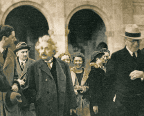 A group of people, including a man with distinctive white hair and a mustache resembling Albert Einstein, walks together outside a large stone building with arched doorways. The mood appears cheerful and informal.