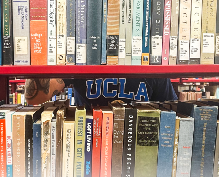 A person wearing a blue UCLA shirt is partially visible behind two rows of assorted books on red shelves in a library or bookstore. Only their chest and part of an arm are visible between the shelves.