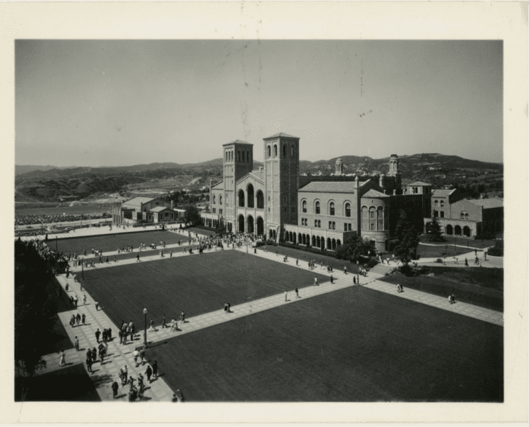 Black and white photo of a large campus quad with people walking on paths, featuring a prominent brick building with two tall towers and mountains visible in the background.