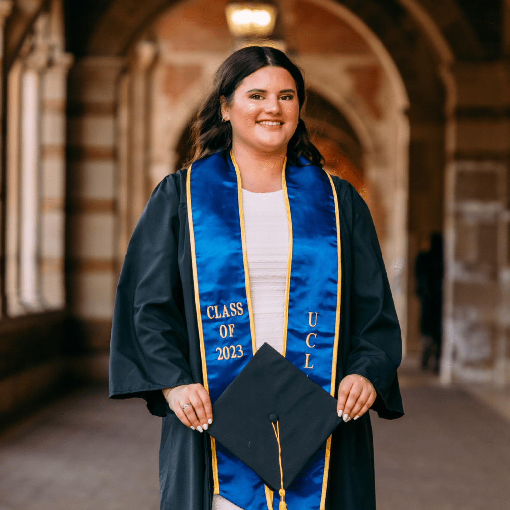 A young woman in a graduation gown and blue stole that reads Class of 2023 UCL stands smiling in an arched hallway, holding a graduation cap.