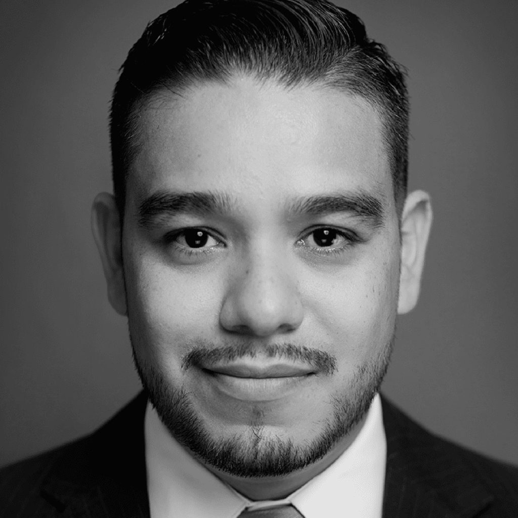Black and white close-up portrait of a man with short dark hair, trimmed beard, and mustache, wearing a suit and tie, smiling slightly, and looking directly at the camera.