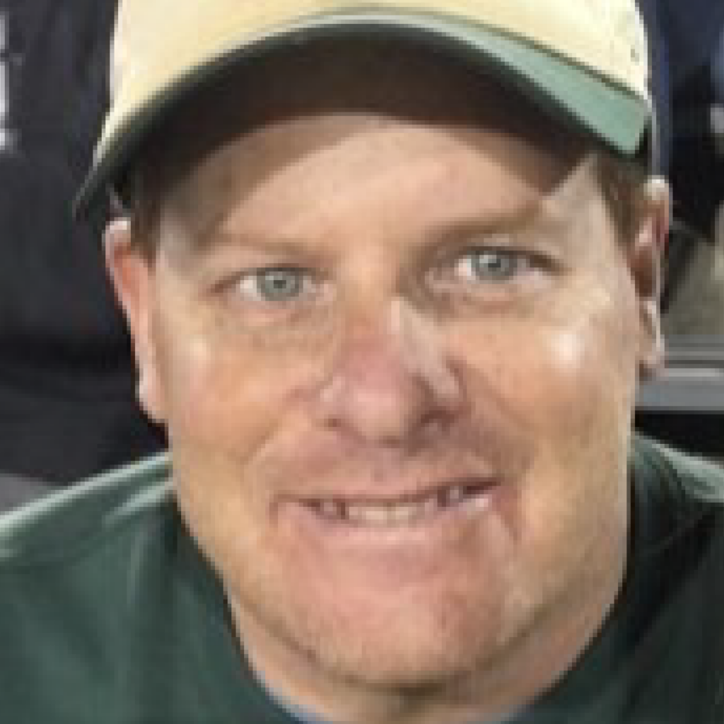 A man wearing a tan and green baseball cap and a green shirt smiles at the camera; his eyes are blue and he has light facial hair.
