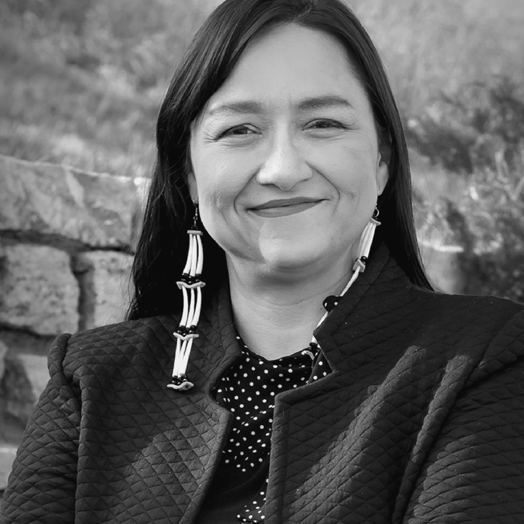 A woman with long dark hair and beaded earrings smiles gently. She wears a textured jacket over a polka dot top. A stone wall and blurred natural background are visible behind her. The image is in black and white.