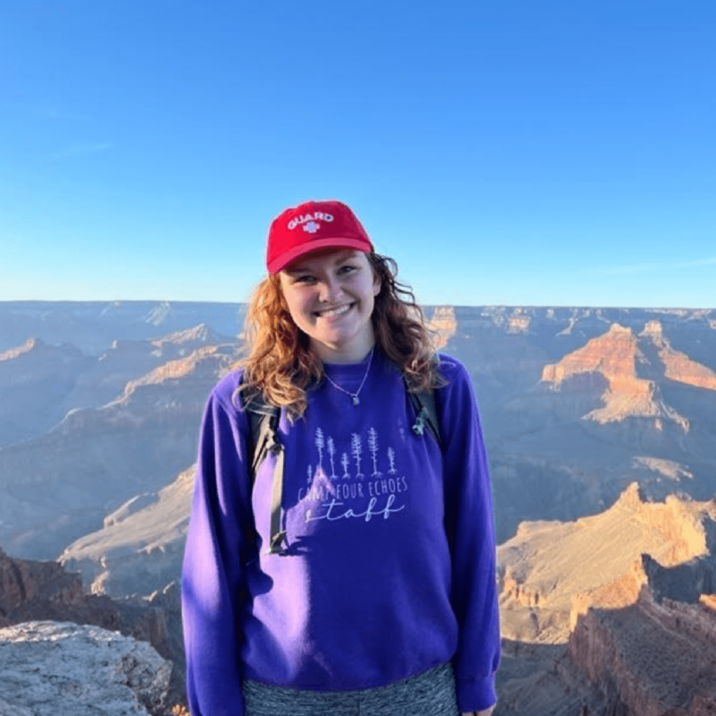 A smiling woman wearing a red cap and purple sweatshirt stands in front of the Grand Canyon under a clear blue sky, with sunlit canyon cliffs in the background.
