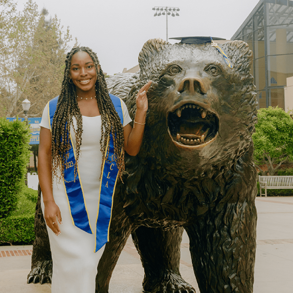 A woman in a white dress and graduation sash stands next to a bear statue wearing a graduation cap, smiling on a university campus.