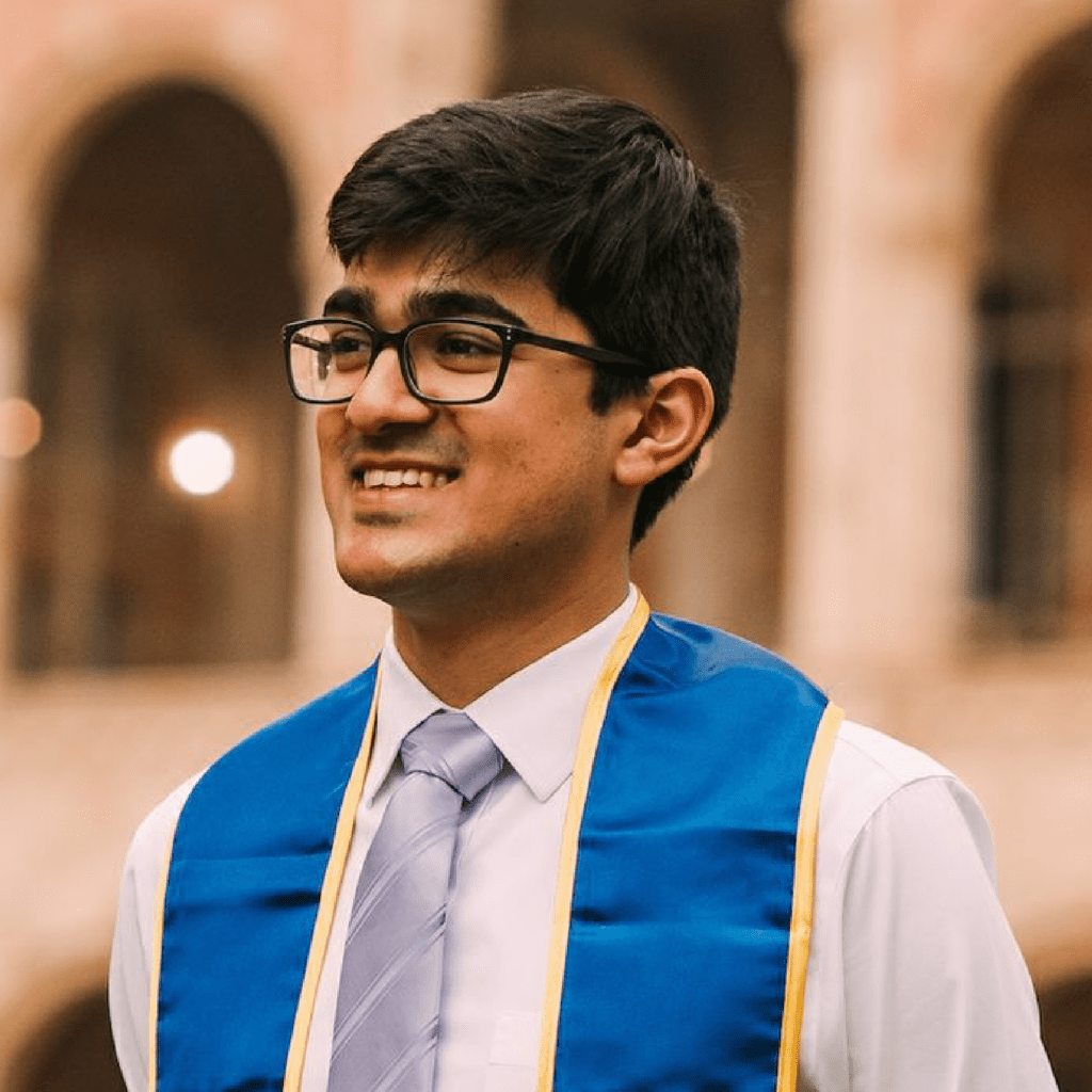 A young man wearing glasses, a white shirt, a tie, and a blue graduation stole smiles while standing outdoors in front of an arched building.