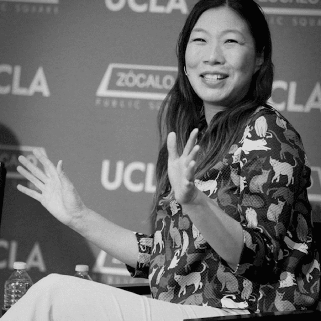 A woman sits and gestures with her hands while speaking at an event. She is smiling and wearing a patterned shirt. Behind her is a backdrop with UCLA and Zócalo Public Square logos.