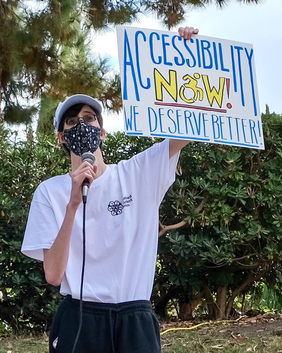A person wearing glasses, a mask, and a white shirt holds a microphone and a sign reading ACCESSIBILITY NOW! WE DESERVE BETTER! while standing outdoors in front of trees and bushes.