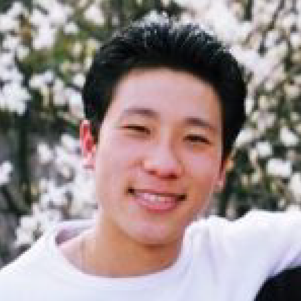 A young person with short black hair smiles at the camera, standing outdoors in front of a background filled with white blooming flowers.