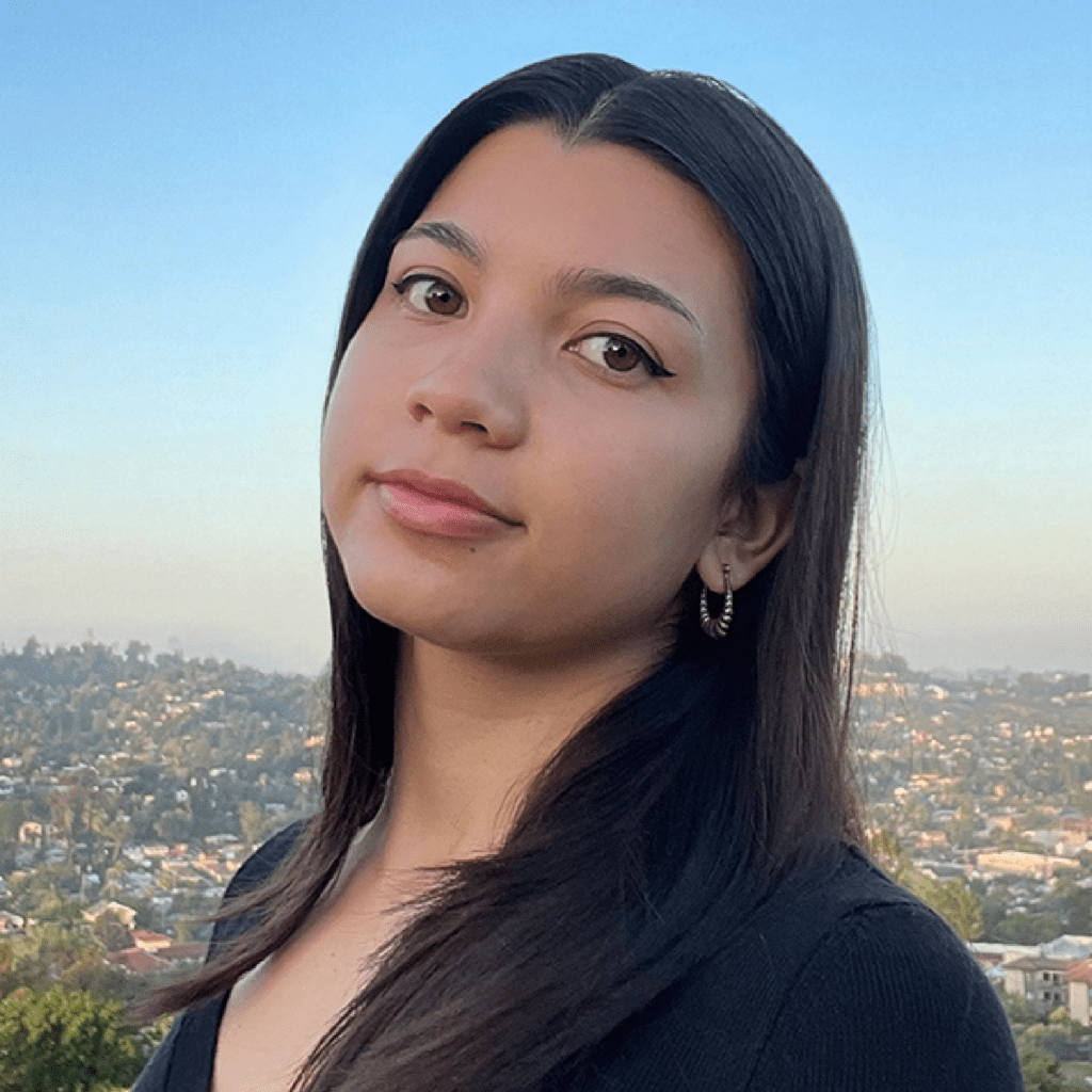 A young woman with long dark hair and hoop earrings stands outdoors, looking at the camera with a neutral expression. A cityscape and clear blue sky are visible in the background.