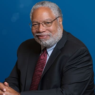 An older man with gray hair and a beard, wearing glasses, a dark suit, light blue shirt, and a red patterned tie, sits against a plain blue background, smiling gently at the camera.