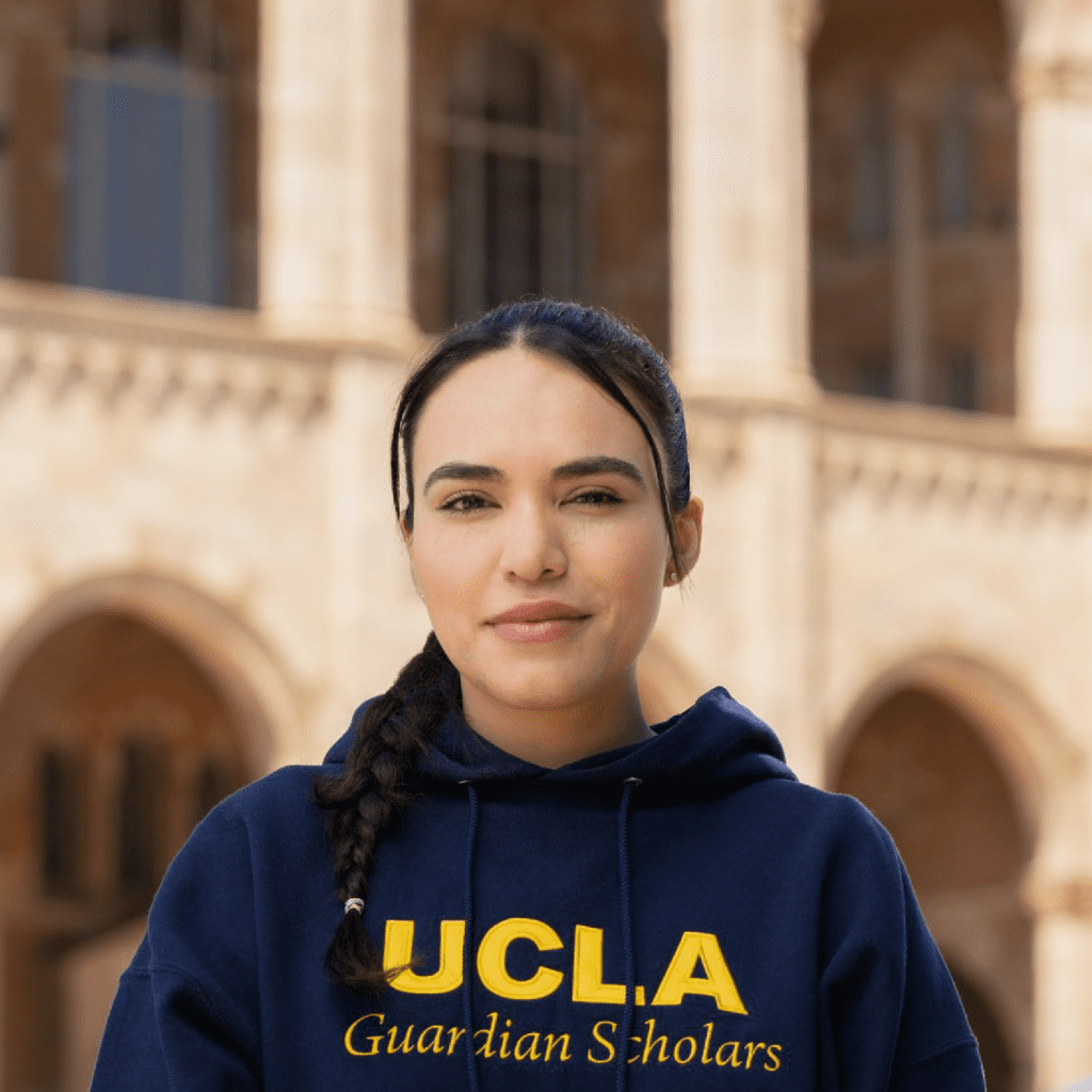 A woman with dark hair in a braid wears a navy blue UCLA Guardian Scholars hoodie and stands in front of a building with arches and columns.