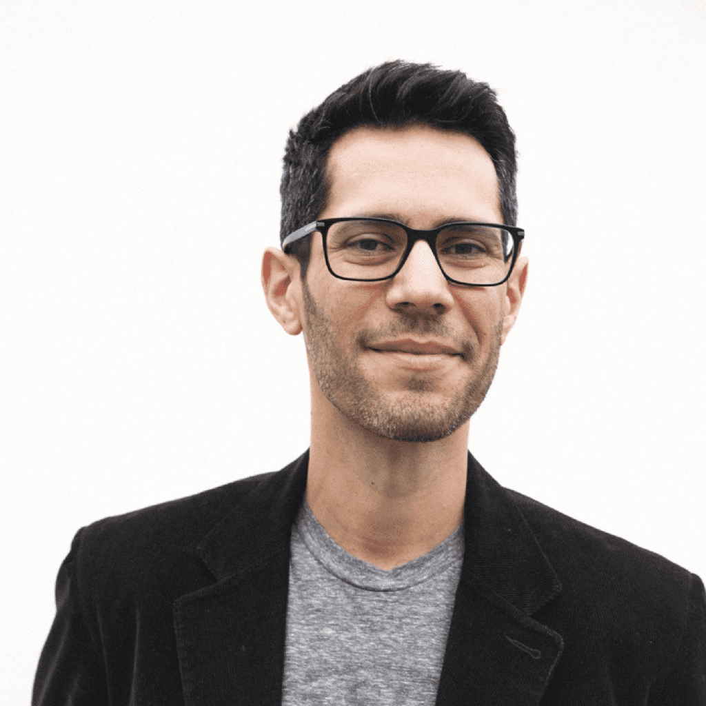 A man with short dark hair, glasses, and light facial hair is smiling slightly. He is wearing a gray t-shirt under a black blazer, standing against a plain white background.