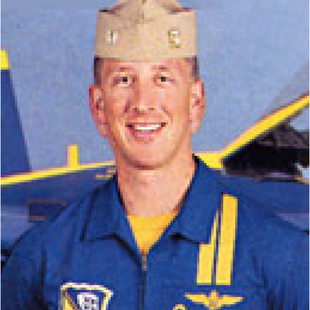 A smiling pilot in a blue and yellow flight suit and tan military cap stands in front of a blue and yellow aircraft.