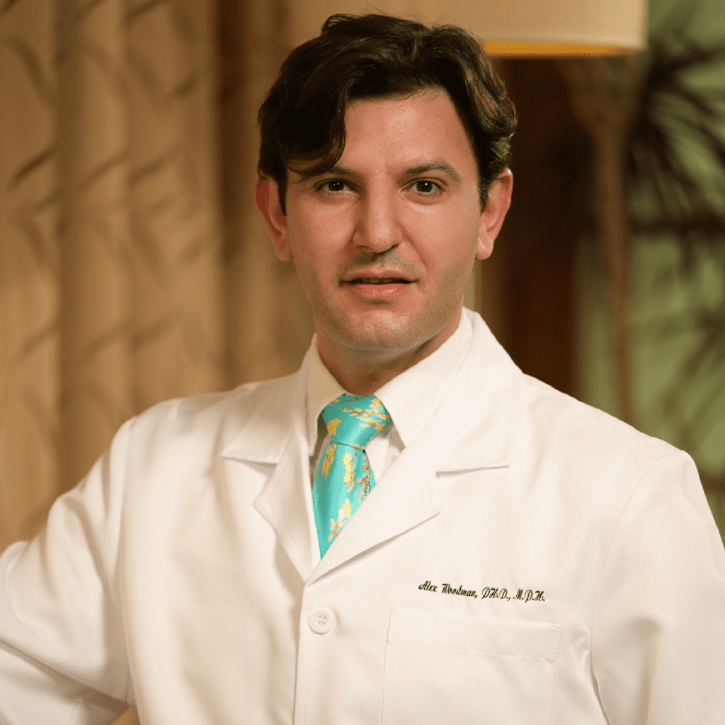 A person with dark hair wearing a white lab coat and a colorful tie stands indoors, looking at the camera with a neutral expression. The background features soft lighting and curtains.