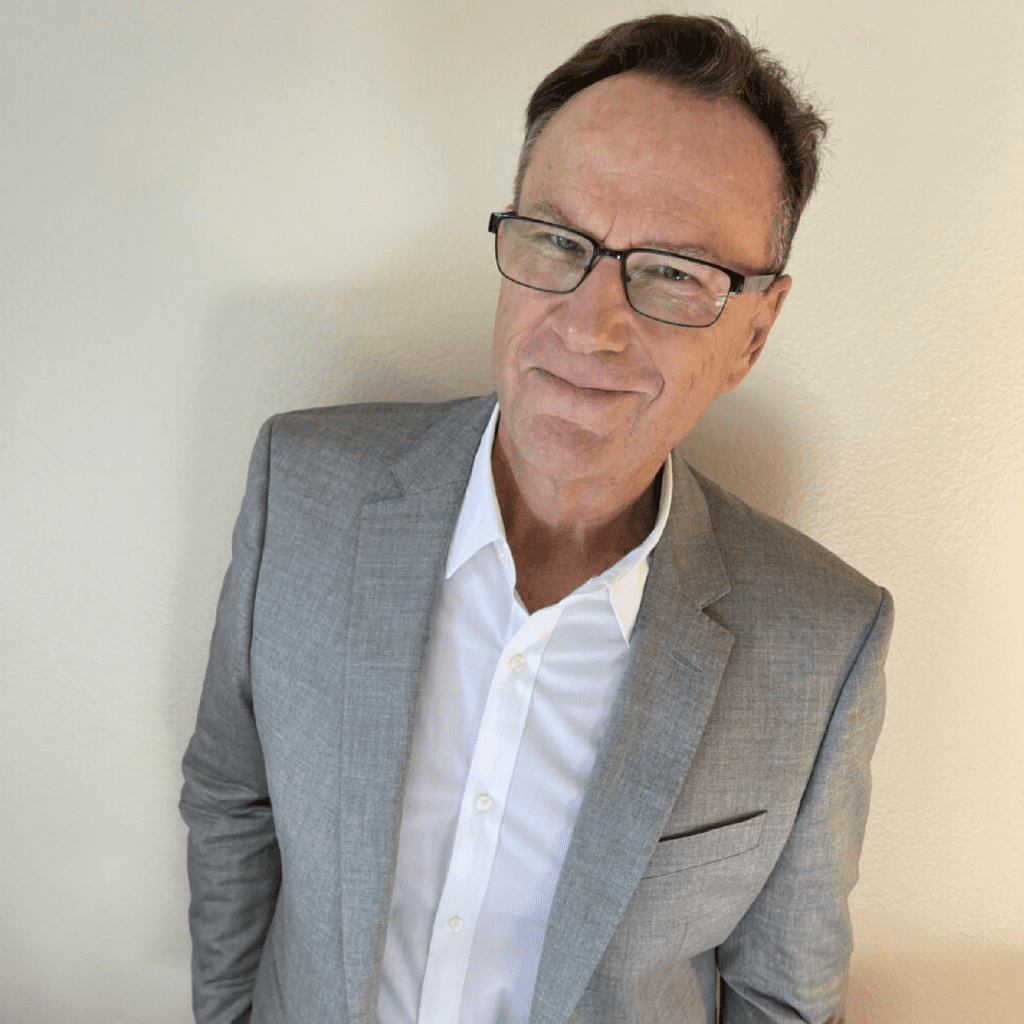 A man wearing glasses and a light gray suit with a white shirt stands against a light-colored wall, smiling slightly at the camera.