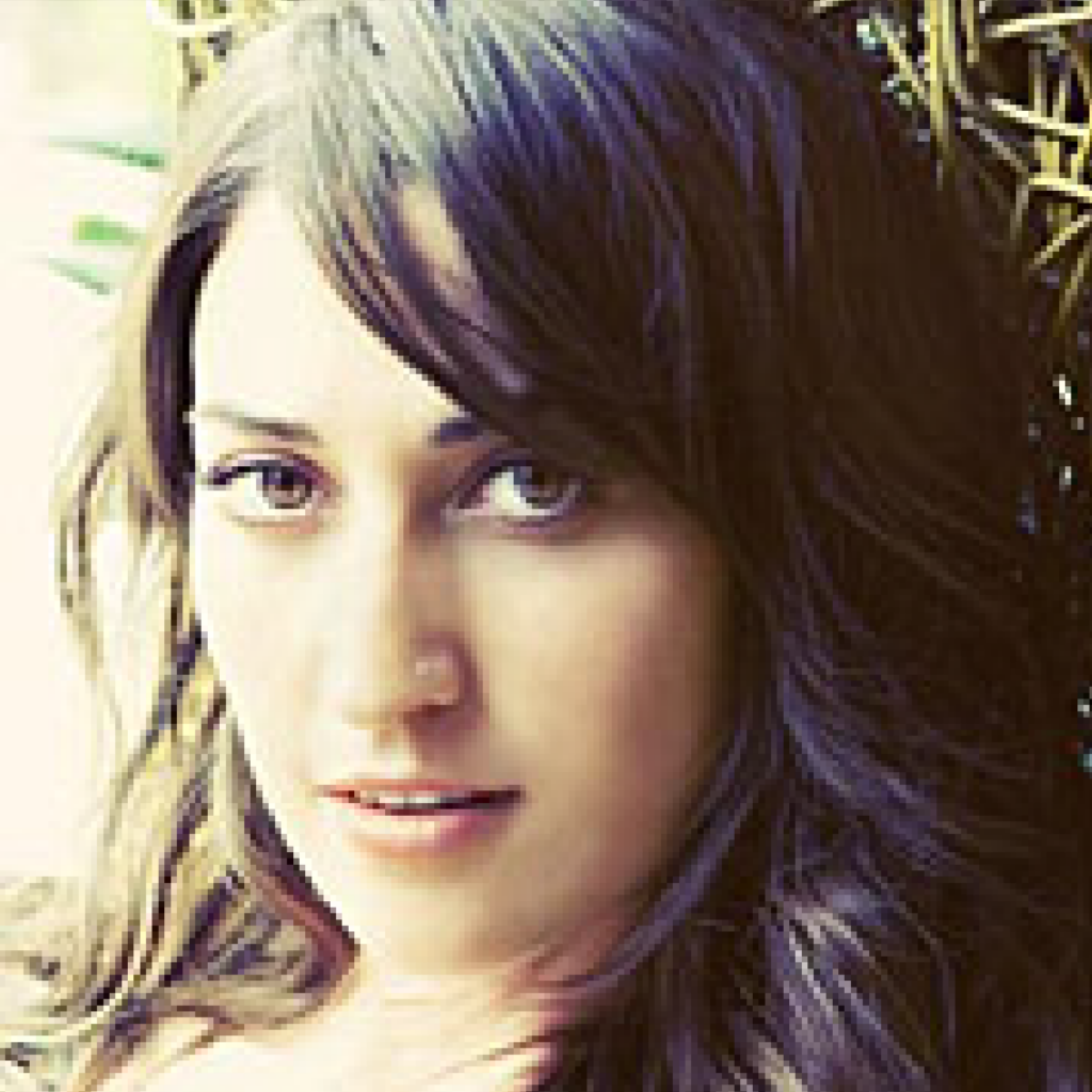 A young woman with long, wavy brown hair and light eyes looks at the camera with a neutral expression, sitting in front of a blurred, natural background.