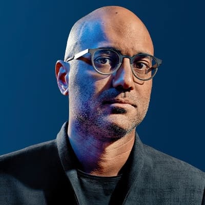 A bald person with glasses and facial stubble wears a dark jacket and black shirt, looking seriously at the camera against a blue gradient background.
