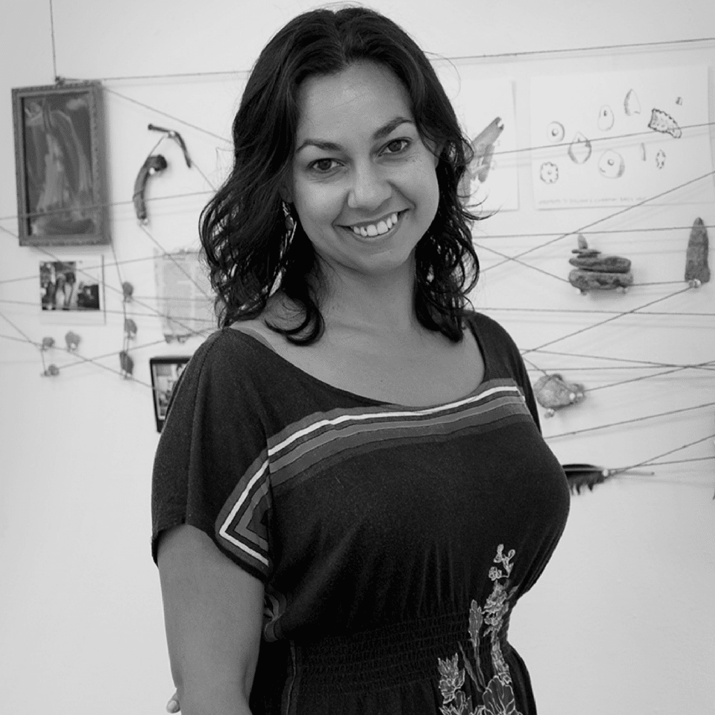 A woman with wavy dark hair, wearing a patterned dress, smiles while standing in front of a wall with art pieces and string installations. The photo is in black and white.