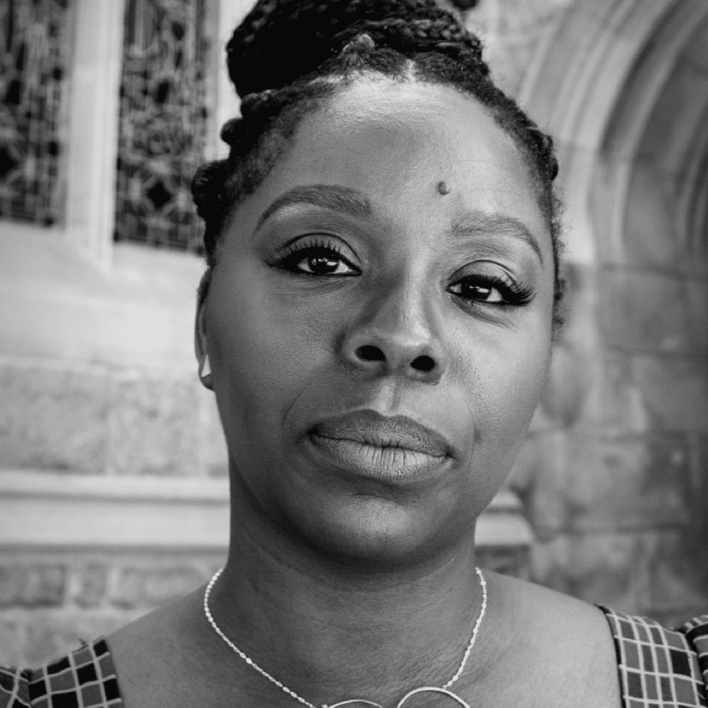 Black and white portrait of a woman with braided hair styled in a bun, wearing a necklace, looking directly at the camera, with a stone building and stained glass window in the background.