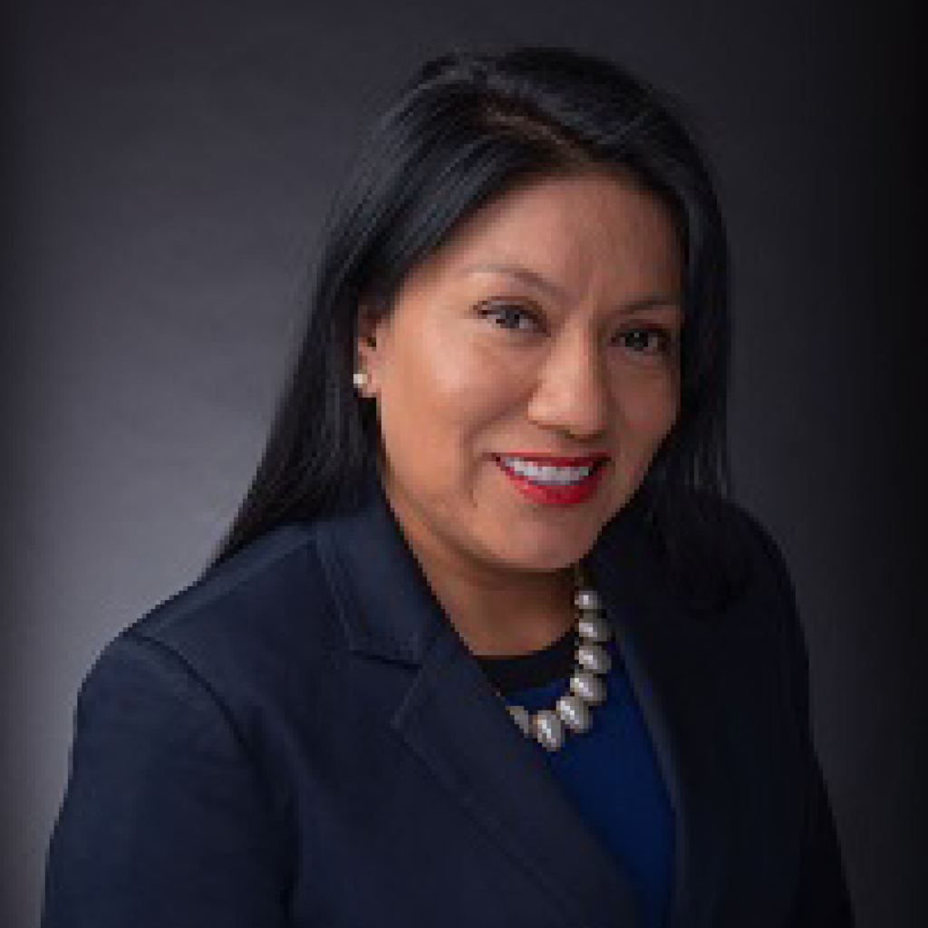 A woman with long black hair, wearing a dark blazer, blue top, pearl necklace, and stud earrings, smiles at the camera against a dark, neutral background.
