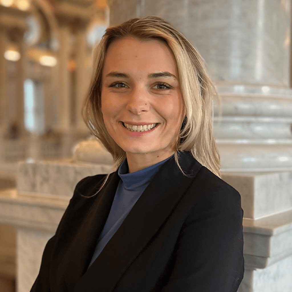A woman with blonde hair wearing a black blazer and blue top smiles while standing indoors in a marble-accented building with blurred background columns.