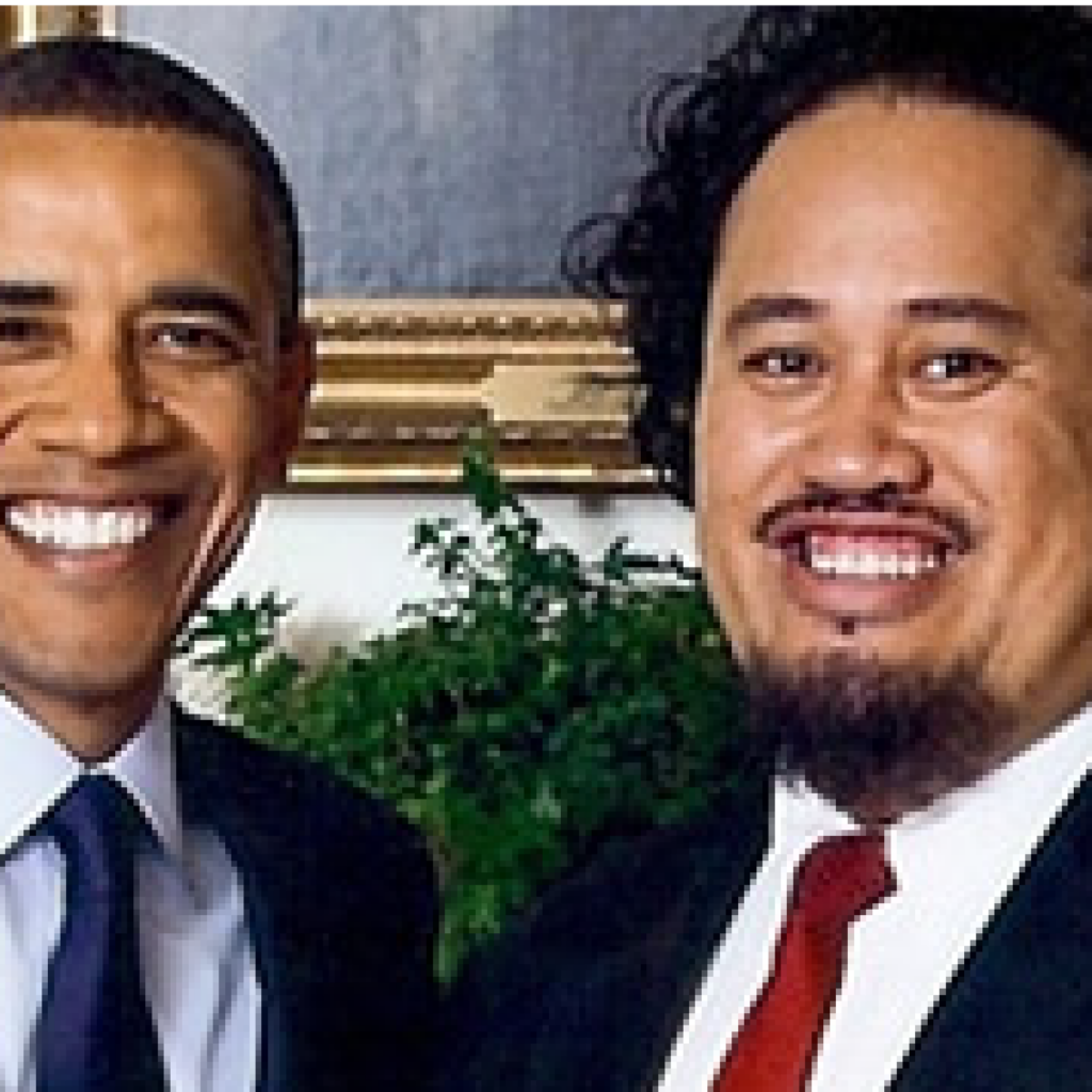 Two men in suits and ties are smiling at the camera while standing close together indoors. There is a framed picture and a green plant in the background.