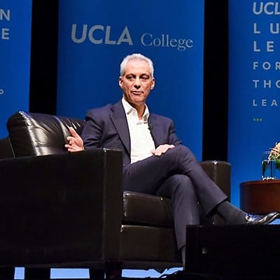A man in a suit sits on a dark armchair, speaking on stage in front of blue banners that read UCLA College and Luskin Lecture for Thought Leadership.