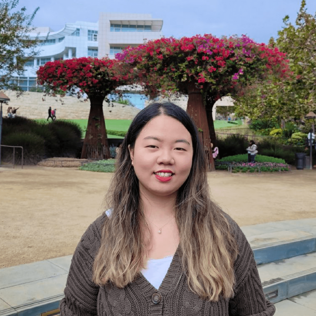 A woman with long, ombre hair stands outdoors, smiling at the camera. Behind her are blooming trees shaped like large planters and a modern building in the background, suggesting a garden or park setting.