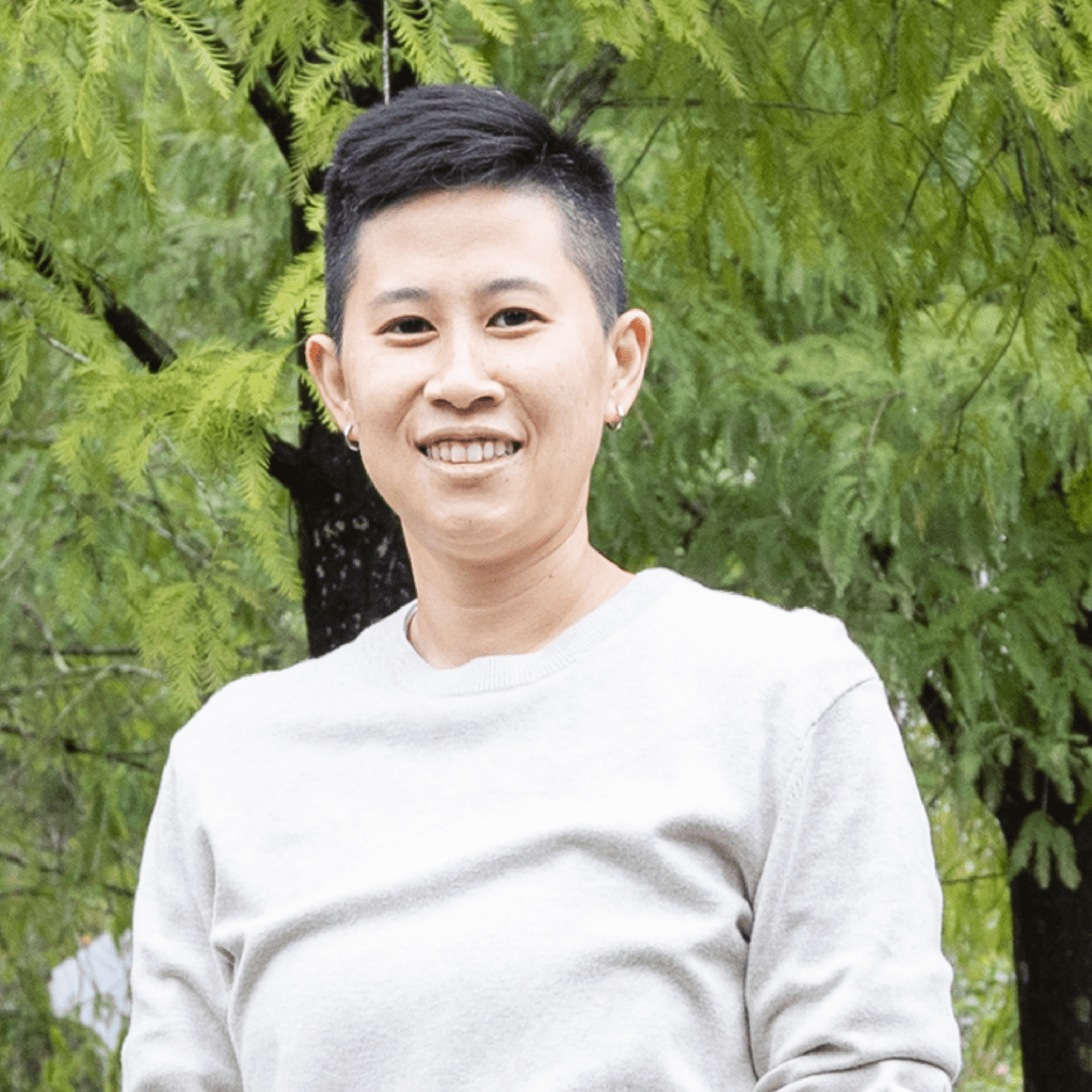 A person with short dark hair, wearing a light sweater, stands outdoors in front of green leafy trees, smiling at the camera.