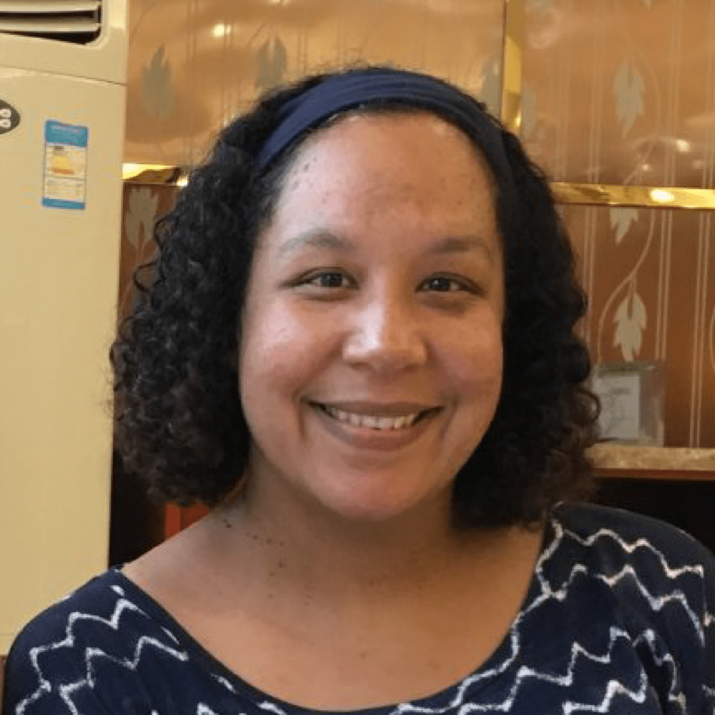 A woman with curly hair, wearing a blue headband and a navy blue patterned top, smiles at the camera. She is indoors with a gold and cream patterned wall and an air conditioner in the background.