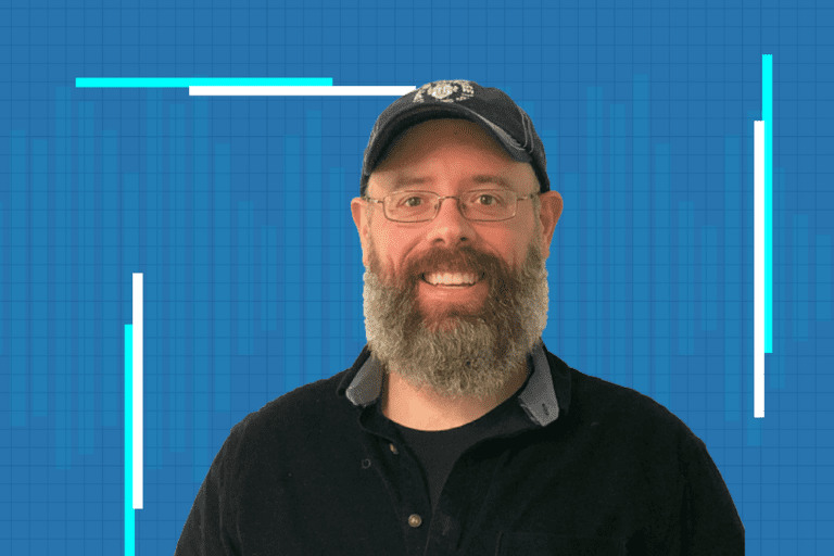 A smiling man with glasses, a full beard, and a dark cap stands in front of a blue background with light blue and white graphic lines.