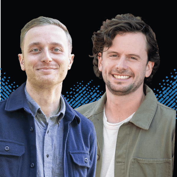 Two men smiling, both wearing jackets over shirts, stand side by side in front of a black background with a blue dotted pattern.
