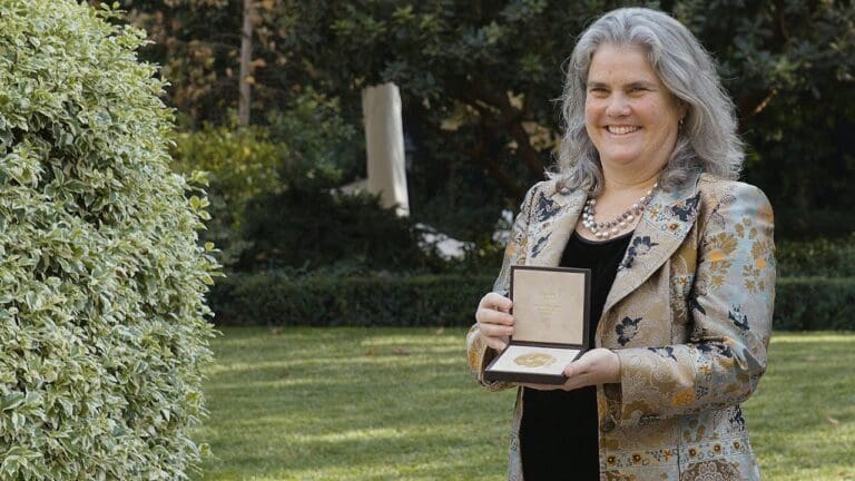 A woman with gray hair, wearing a patterned jacket and pearl necklace, stands outdoors on grass, smiling and holding an open award box with a gold medal inside. Shrubs and trees are in the background.