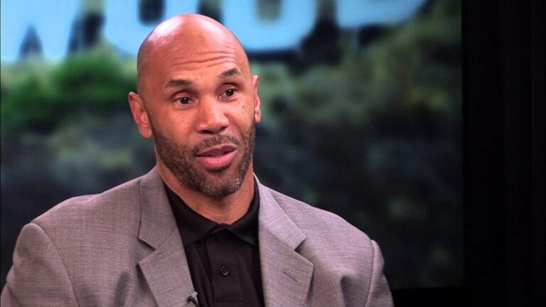 A man with a bald head, trimmed beard, and mustache wears a grey suit jacket over a black shirt, speaking in front of a blurred background.