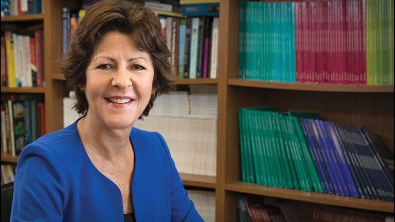 A woman with short brown hair, wearing a blue blazer, smiles while sitting at a desk in front of bookshelves filled with colorful books and journals.