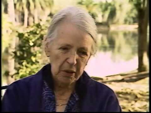 An elderly woman with gray hair pulled back, wearing a dark blue jacket, sits outdoors near a pond with trees and greenery in the background. She has a calm, thoughtful expression.