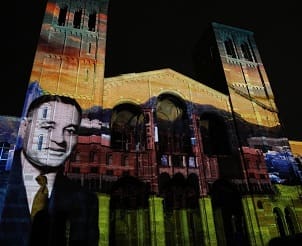 A nighttime image of a building with twin towers illuminated by colorful light projections, including the large projected image of a smiling man in a suit on the left side of the facade.